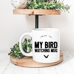 May include: White ceramic mug with black text that reads "This is my bird watching mug" and a black silhouette of a bird. The mug is on a wooden tiered stand with green plants.