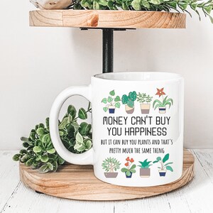 Puede incluir: Taza de cer&aacute;mica blanca con asa negra. La taza tiene un gr&aacute;fico de plantas en maceta y el texto "MONEY CAN'T BUY YOU HAPPINESS BUT IT CAN BUY YOU PLANTS AND THAT'S PRETTY MUCH THE SAME THING".
