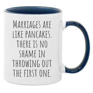 May include: White ceramic coffee mug with a blue rim and handle. The mug has a funny saying printed on it: "Marriages are like pancakes. There is no shame in throwing out the first one."