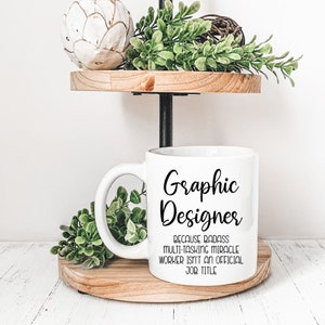 May include: White ceramic coffee mug with a black graphic that reads "Graphic Designer because badass multi-tasking miracle worker isn't an official job title."