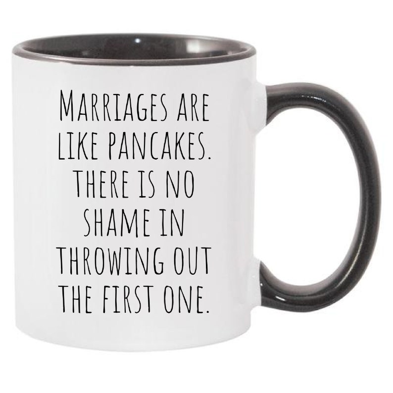 May include: White ceramic coffee mug with a black rim and handle. The mug has a funny saying printed on it: "Marriages are like pancakes. There is no shame in throwing out the first one."