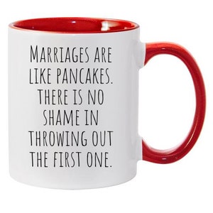May include: White ceramic coffee mug with a red rim and handle. The mug has a funny saying printed on it: "Marriages are like pancakes. There is no shame in throwing out the first one."
