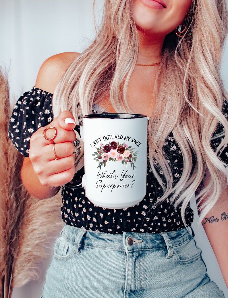 Pu&ograve; includere: Una tazza da caff&egrave; bianca con bordo e manico neri. La tazza ha un motivo floreale e il testo "I just outlived my knee. What's your superpower?"