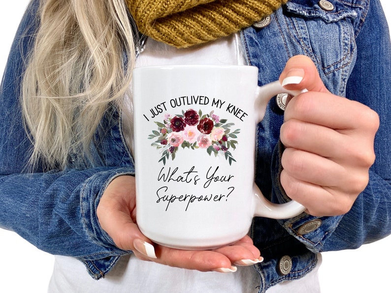 Pu&ograve; includere: Una tazza in ceramica bianca con un motivo floreale e il testo "I just outlived my knee. What's your superpower?"