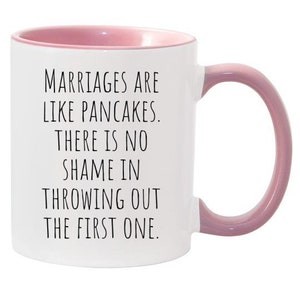 May include: White ceramic coffee mug with a pink rim and handle. The mug has a funny saying printed on it: "Marriages are like pancakes. There is no shame in throwing out the first one."