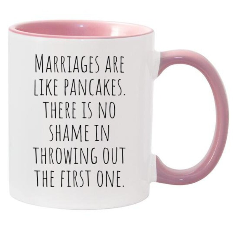 May include: White ceramic coffee mug with a pink rim and handle. The mug has a funny saying printed on it: "Marriages are like pancakes. There is no shame in throwing out the first one."