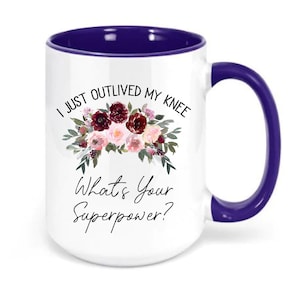 Pu&ograve; includere: Una tazza da caff&egrave; bianca con bordo e manico viola. La tazza presenta un motivo floreale e il testo "I just outlived my knee. What's your superpower?"