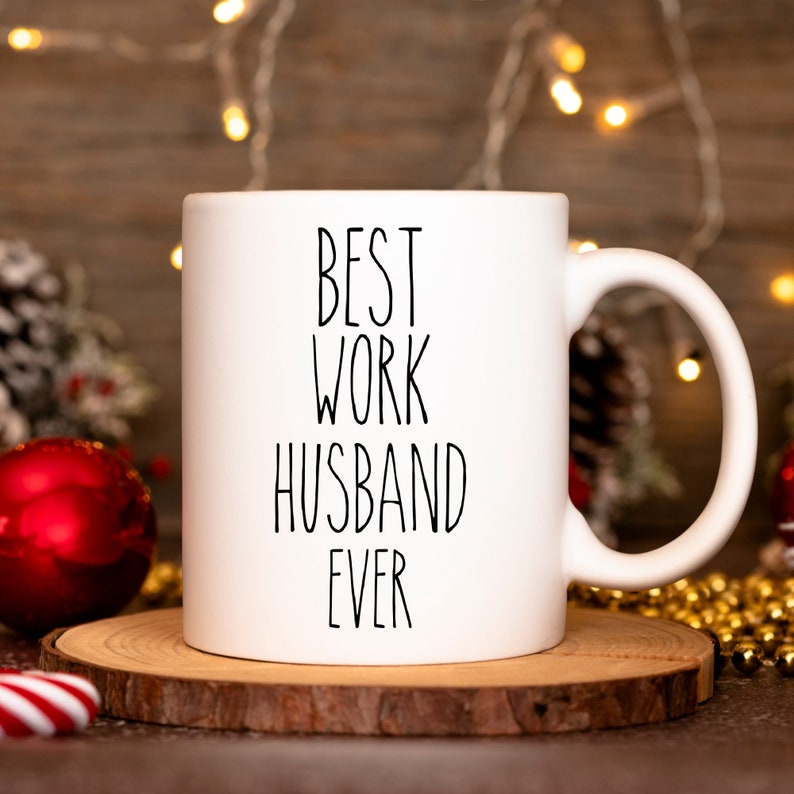 May include: White ceramic coffee mug with black text that reads "Best Work Husband Ever". The mug is sitting on a wooden coaster with gold beads and a red ornament in the background.