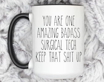 Puede incluir: Taza de cerámica blanca con asa y borde negros. La taza tiene el texto "You are one amazing badass surgical tech keep that shit up" impreso en negro.