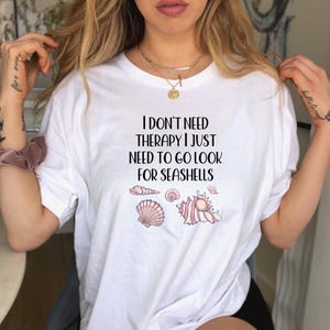 May include: White t-shirt featuring the text "I DON'T NEED THERAPY I JUST NEED TO GO LOOK FOR SEASHELLS" and seashell illustrations in pink and beige. The shirt has a crew neck and short sleeves.