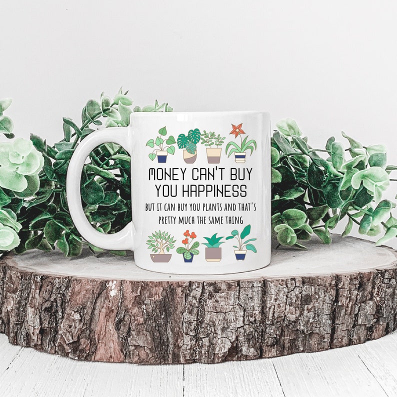 Puede incluir: Taza de cer&aacute;mica blanca con una ilustraci&oacute;n en negro y verde de plantas y el texto "MONEY CAN'T BUY YOU HAPPINESS BUT IT CAN BUY YOU PLANTS AND THAT'S PRETTY MUCH THE SAME THING".