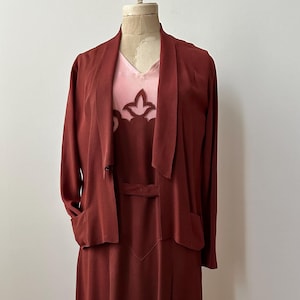 Op de afbeelding: Een maronkleurige jurk met een bijpassend jasje. De jurk heeft een V-hals en een roze onderlaag met een uitgesneden ontwerp. Het jasje heeft een shawlkraag en lange mouwen.