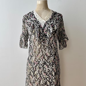Op de afbeelding: Een vintage zwarte jurk met een witte gestippelde halslijn en korte mouwen. De jurk heeft een doorschijnende overlay met een patroon van witte, roze en gele penseelstreken. De jurk wordt op een mannequin getoond.