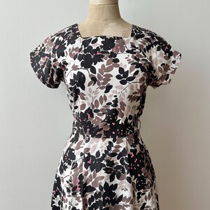 Op de afbeelding: Een vintage jurk met een vierkante halslijn en korte mouwen. De jurk heeft een bloemenpatroon met zwarte, bruine en roze bladeren op een witte achtergrond. De jurk heeft een getailleerd lijfje en een uitlopende rok.