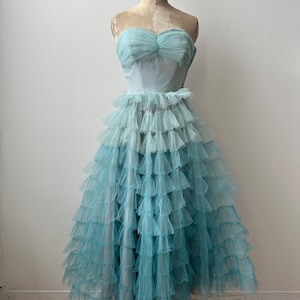 Op de afbeelding: Een vintage, strapless avondjurk in blauwtinten. De lijfje is lichtblauw, terwijl de rok lagen gerimpelde tule heeft in een donkerdere, blauwgroene tint. De jurk wordt op een mannequin getoond.