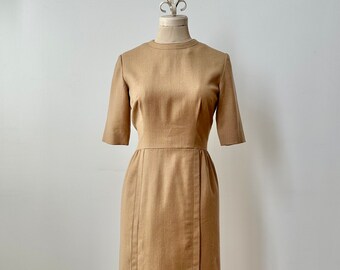 Puede incluir: Vestido color beige, largo hasta la rodilla, con mangas cortas y cuello redondo. El vestido presenta un corpiño entallado y una falda recta con pliegues verticales. El vestido se exhibe en un maniquí.