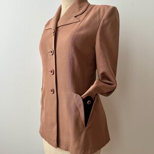 May include: A vintage-style, dusty rose blazer with a classic collar and three matching buttons. The blazer features two front pockets and long sleeves. The garment is displayed on a mannequin, showcasing its tailored fit and design.