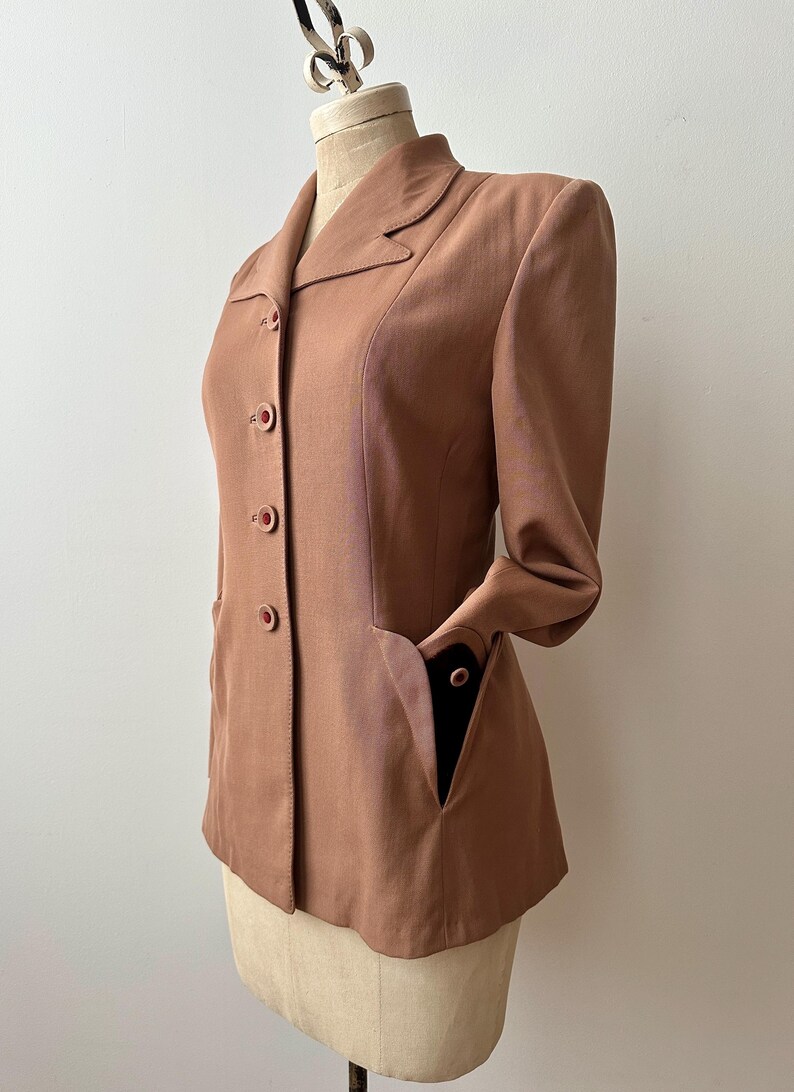 May include: A vintage-style, dusty rose blazer with a classic collar and three matching buttons. The blazer features two front pockets and long sleeves. The garment is displayed on a mannequin, showcasing its tailored fit and design.
