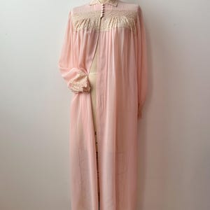 Op de afbeelding: Een vintage, lichtroze badjas met lange mouwen. De badjas heeft een kraag, delicate kanten afwerking en een knoopsluiting aan de voorkant. De badjas wordt op een mannequin gepresenteerd.