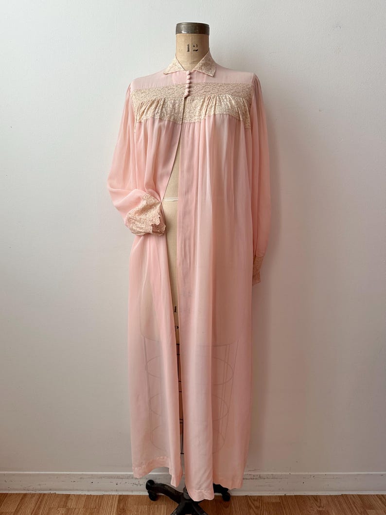 Op de afbeelding: Een vintage, lichtroze badjas met lange mouwen. De badjas heeft een kraag, delicate kanten afwerking en een knoopsluiting aan de voorkant. De badjas wordt op een mannequin gepresenteerd.
