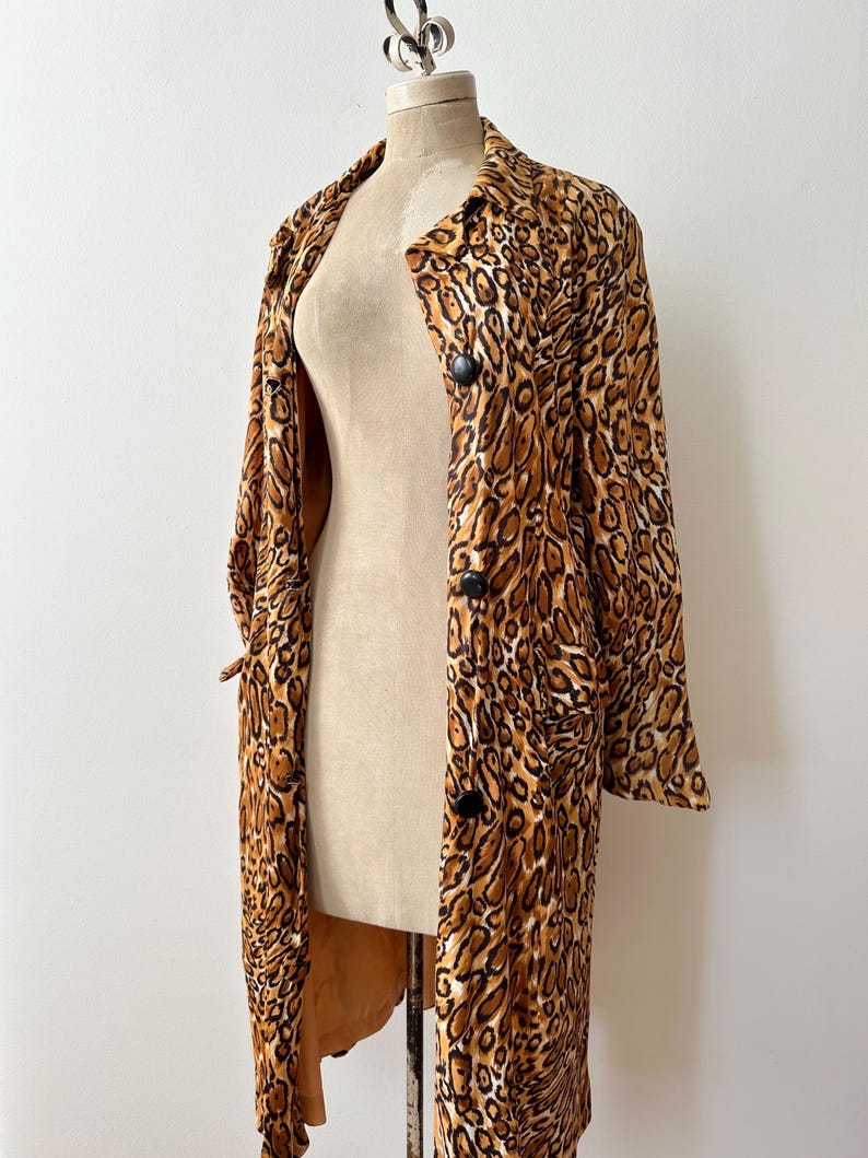 Puede incluir: Un abrigo largo con estampado de leopardo, botones negros y cuello. El abrigo se exhibe en un maniqu&iacute; sobre un fondo blanco. El patr&oacute;n del abrigo presenta manchas marrones y beige.