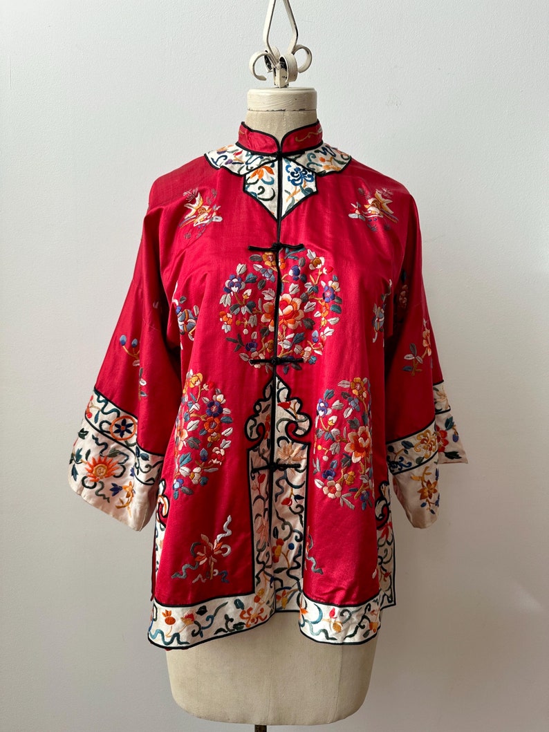 May include: A red silk Chinese jacket with intricate floral embroidery in shades of blue, green, and yellow. The jacket has a mandarin collar and a front closure with decorative buttons.