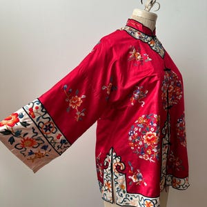 May include: A red silk jacket with intricate floral embroidery in shades of pink, blue, and yellow. The jacket has a mandarin collar and long sleeves with decorative trim.