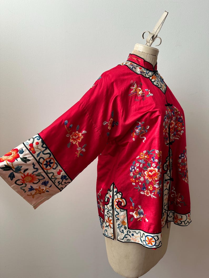 May include: A red silk jacket with intricate floral embroidery in shades of pink, blue, and yellow. The jacket has a mandarin collar and long sleeves with decorative trim.