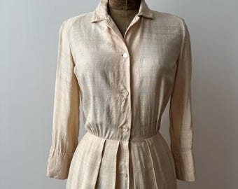 May include: A light beige vintage dress with long sleeves and a button-down front. The dress features a collared neckline and a pleated skirt. The dress is displayed on a mannequin.