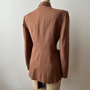 May include: A brown blazer displayed on a mannequin. The tailored jacket features a notched collar, long sleeves, and a fitted silhouette. The back view highlights the structured design and vertical seams. The blazer is a solid, warm brown color.