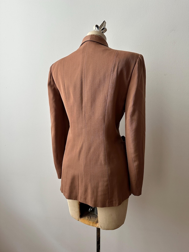 May include: A brown blazer displayed on a mannequin. The tailored jacket features a notched collar, long sleeves, and a fitted silhouette. The back view highlights the structured design and vertical seams. The blazer is a solid, warm brown color.
