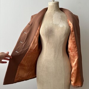 May include: A brown blazer displayed on a vintage dress form. The blazer is open, revealing a satin-like orange lining. The garment has three buttons and a classic lapel design. The dress form is a neutral beige color.