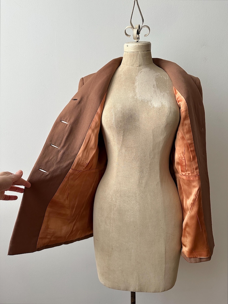 May include: A brown blazer displayed on a vintage dress form. The blazer is open, revealing a satin-like orange lining. The garment has three buttons and a classic lapel design. The dress form is a neutral beige color.