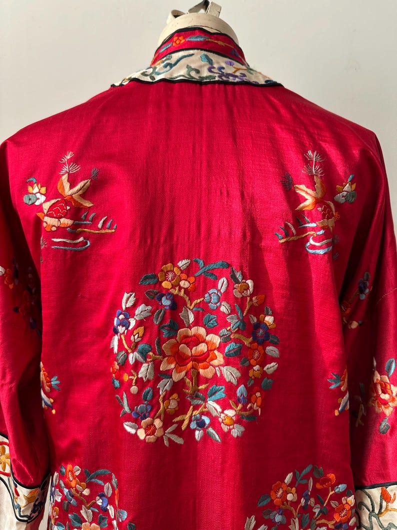 May include: A red silk robe with intricate floral embroidery in shades of blue, green, orange, and yellow. The robe has a mandarin collar and is adorned with birds and flowers.