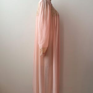 Op de afbeelding: Een vintage, lichtroze nachtjapon met lange mouwen en een zoom tot op de grond. De nachtjapon heeft delicate kanten details op het lijfje en de manchetten. Het kledingstuk wordt op een mannequin gepresenteerd.