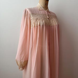 Op de afbeelding: Een vintage, lichtroze nachtjapon met lange mouwen en een kraag. De nachtjapon heeft een beige kanten paneel over de borst en bij de manchetten. Het kledingstuk wordt op een mannequin getoond.