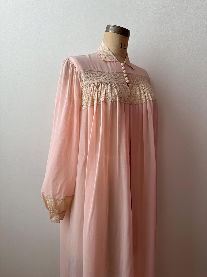 Op de afbeelding: Een vintage, lichtroze nachtjapon met lange mouwen en een kraag. De nachtjapon heeft een beige kanten paneel over de borst en bij de manchetten. Het kledingstuk wordt op een mannequin getoond.