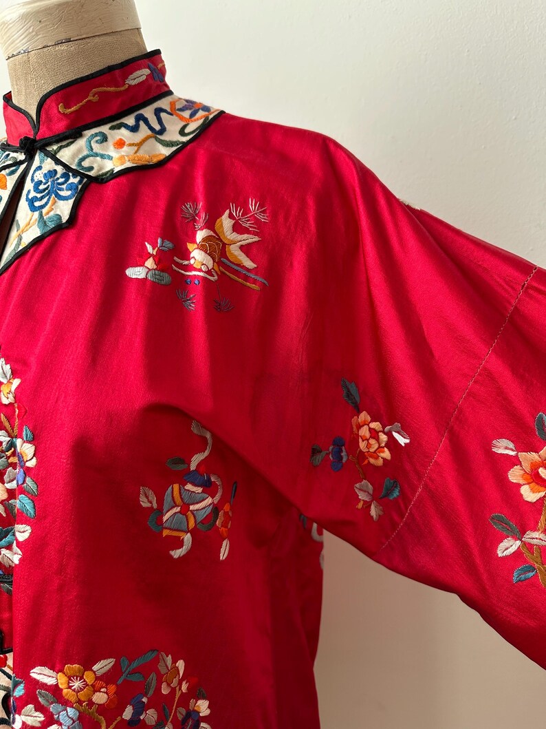 May include: A red silk robe with intricate floral embroidery in shades of blue, green, yellow, and pink. The robe has a mandarin collar with a black trim and a white and blue embroidered design.