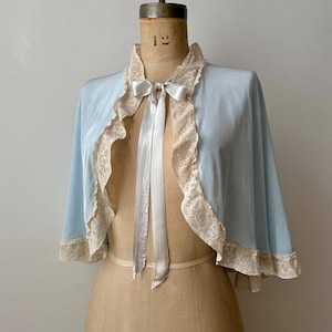 May include: A light blue capelet featuring a ruffled cream-coloured lace trim along the front edges and hem. The garment is secured at the neckline with a matching light blue satin ribbon tied into a bow, creating a soft, draped silhouette.