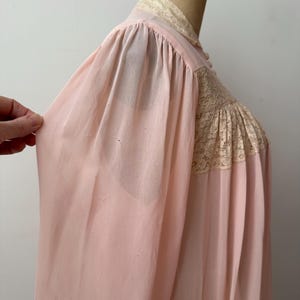 Op de afbeelding: Zachtroze vintage nachtjapon met kanten details op het lijfje en de kraag. De nachtjapon heeft lange mouwen en een vloeiend silhouet. De stof lijkt een lichtgewicht, semi-transparant materiaal te zijn.