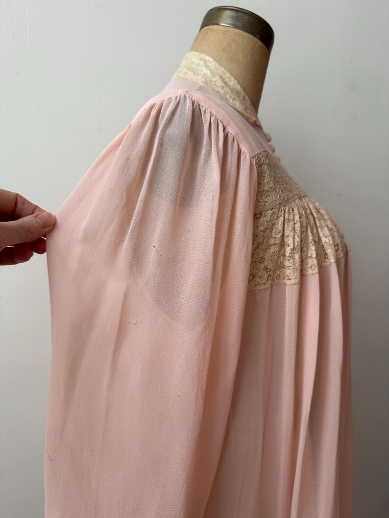 Op de afbeelding: Zachtroze vintage nachtjapon met kanten details op het lijfje en de kraag. De nachtjapon heeft lange mouwen en een vloeiend silhouet. De stof lijkt een lichtgewicht, semi-transparant materiaal te zijn.