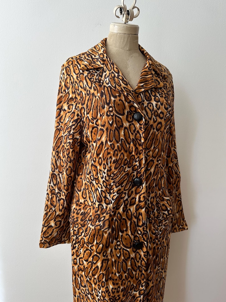 Puede incluir: Un abrigo de manga larga con estampado de leopardo en tonos marr&oacute;n y beige. El abrigo tiene cuello y botones negros. La prenda se exhibe en un maniqu&iacute;. Se aprecia la longitud del abrigo.