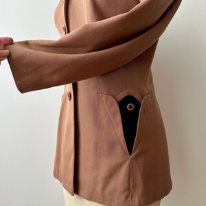 May include: A tailored, light brown blazer with a notched collar and long sleeves. The blazer features a unique pocket design with a black interior and a single button closure. The garment is displayed on a mannequin.