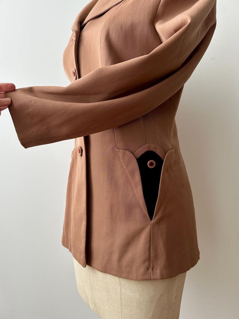 May include: A tailored, light brown blazer with a notched collar and long sleeves. The blazer features a unique pocket design with a black interior and a single button closure. The garment is displayed on a mannequin.