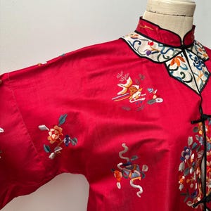 May include: A red silk Chinese-style jacket with intricate floral embroidery in shades of blue, green, yellow, and orange. The jacket has a mandarin collar and a button closure.