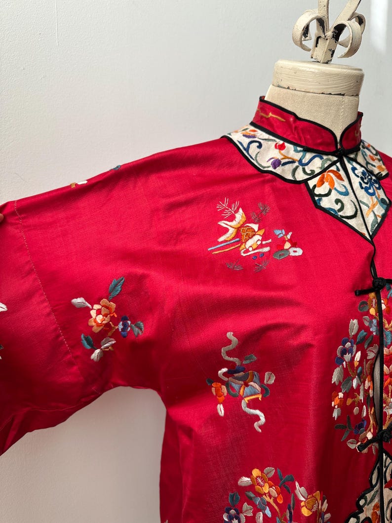 May include: A red silk Chinese-style jacket with intricate floral embroidery in shades of blue, green, yellow, and orange. The jacket has a mandarin collar and a button closure.