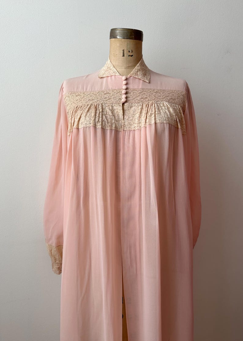 Op de afbeelding: Een vintage, lichtroze nachtjapon met lange mouwen en een kraag. De buste heeft een decoratief kanten paneel met een bloemmotief. De nachtjapon wordt op een mannequin getoond. De stof lijkt licht en vloeiend.