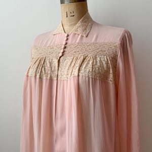 Op de afbeelding: Een vintage, lichtroze nachtjapon met een kraag en lange mouwen. De bodice heeft een beige kanten paneel en kleine, bijpassende knopen. De transparante stof valt naar beneden en cre&euml;ert een vloeiend silhouet. De nachtjapon wordt op een mannequin getoond.