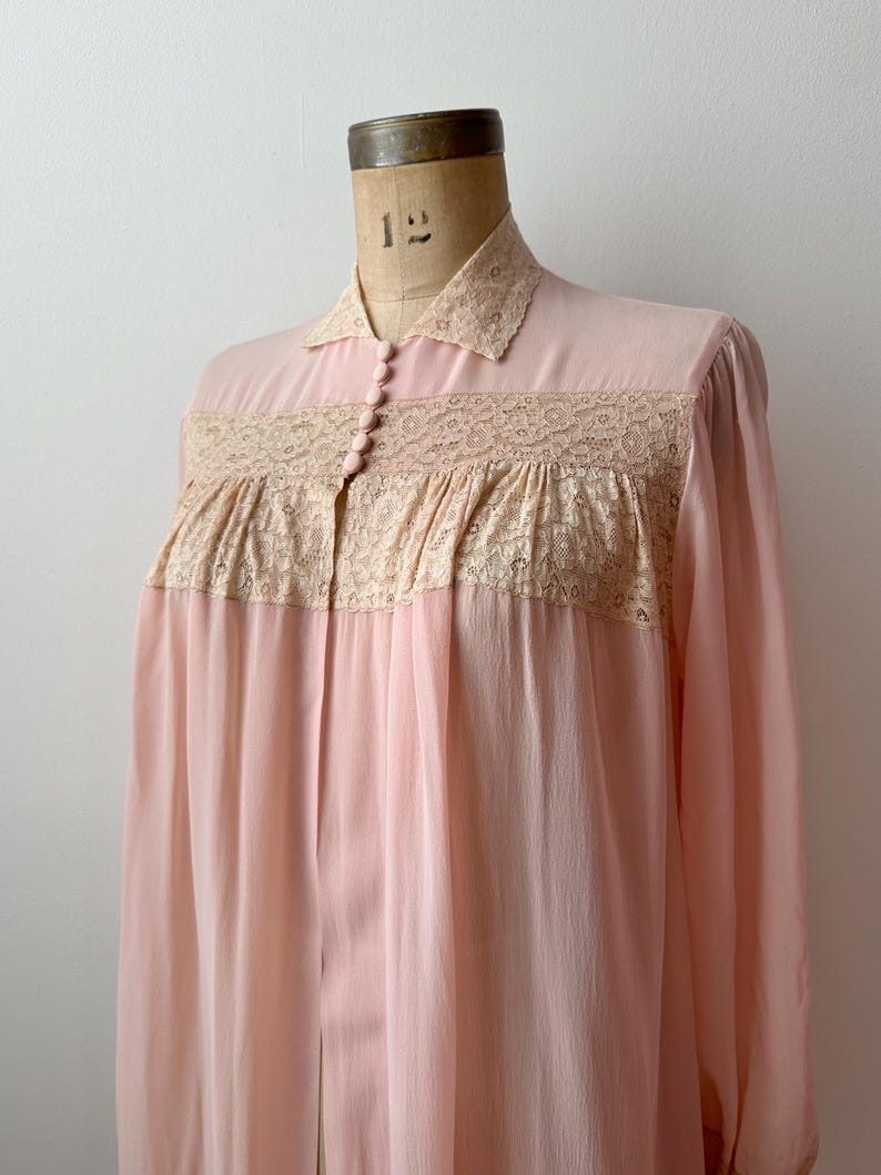 Op de afbeelding: Een vintage, lichtroze nachtjapon met een kraag en lange mouwen. De bodice heeft een beige kanten paneel en kleine, bijpassende knopen. De transparante stof valt naar beneden en cre&euml;ert een vloeiend silhouet. De nachtjapon wordt op een mannequin getoond.