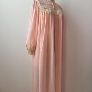 Op de afbeelding: Een vintage, lichtroze nachtjapon met een lengte tot op de grond. De japon heeft lange mouwen met gouden kanten manchetten, een hoge halslijn en een lijfje met gouden kanten details. De japon wordt op een mannequin getoond.
